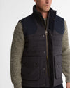 A man wearing a dark navy quilted vest over a textured olive-green knit sweater, shown from the chest up against a light gray studio background.