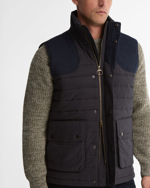A man wearing a dark navy quilted vest over a textured olive-green knit sweater, shown from the chest up against a light gray studio background.