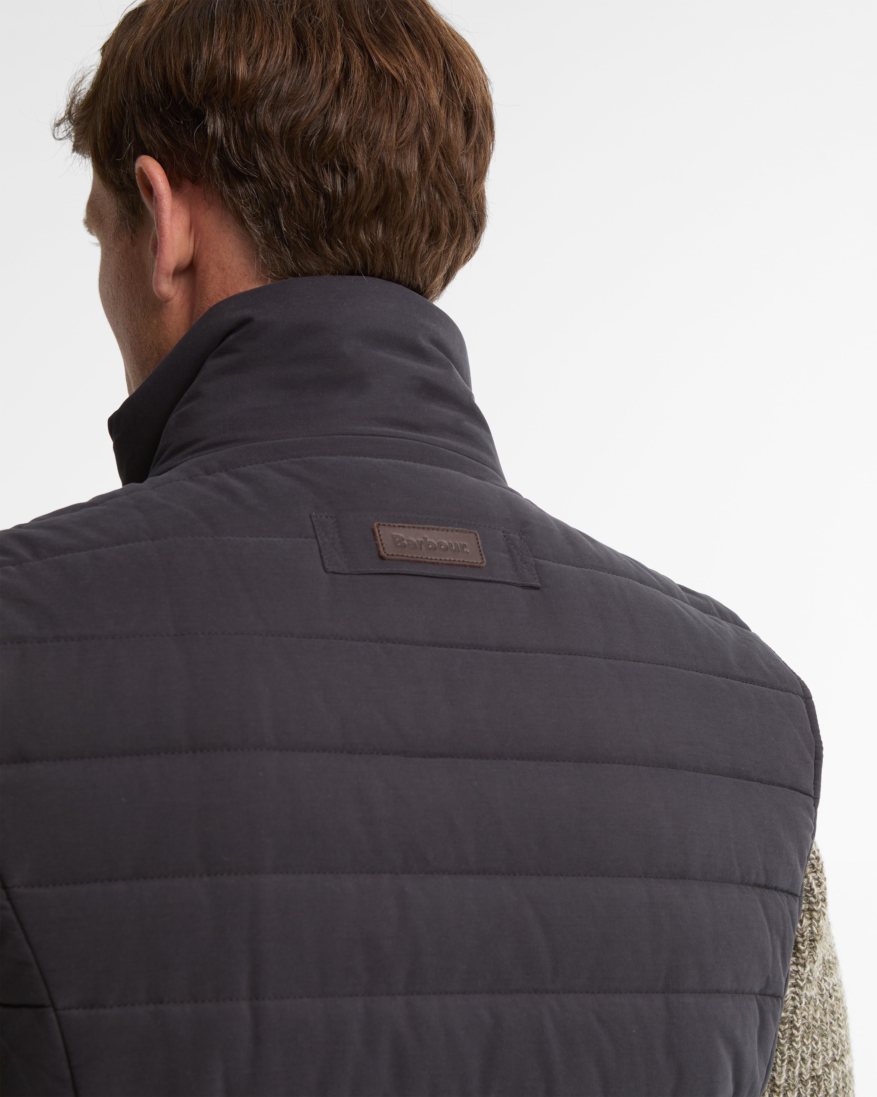 Close-up back view of a man wearing a dark navy quilted Barbour puffer vest, showing the brand patch on the upper back against a light gray background.