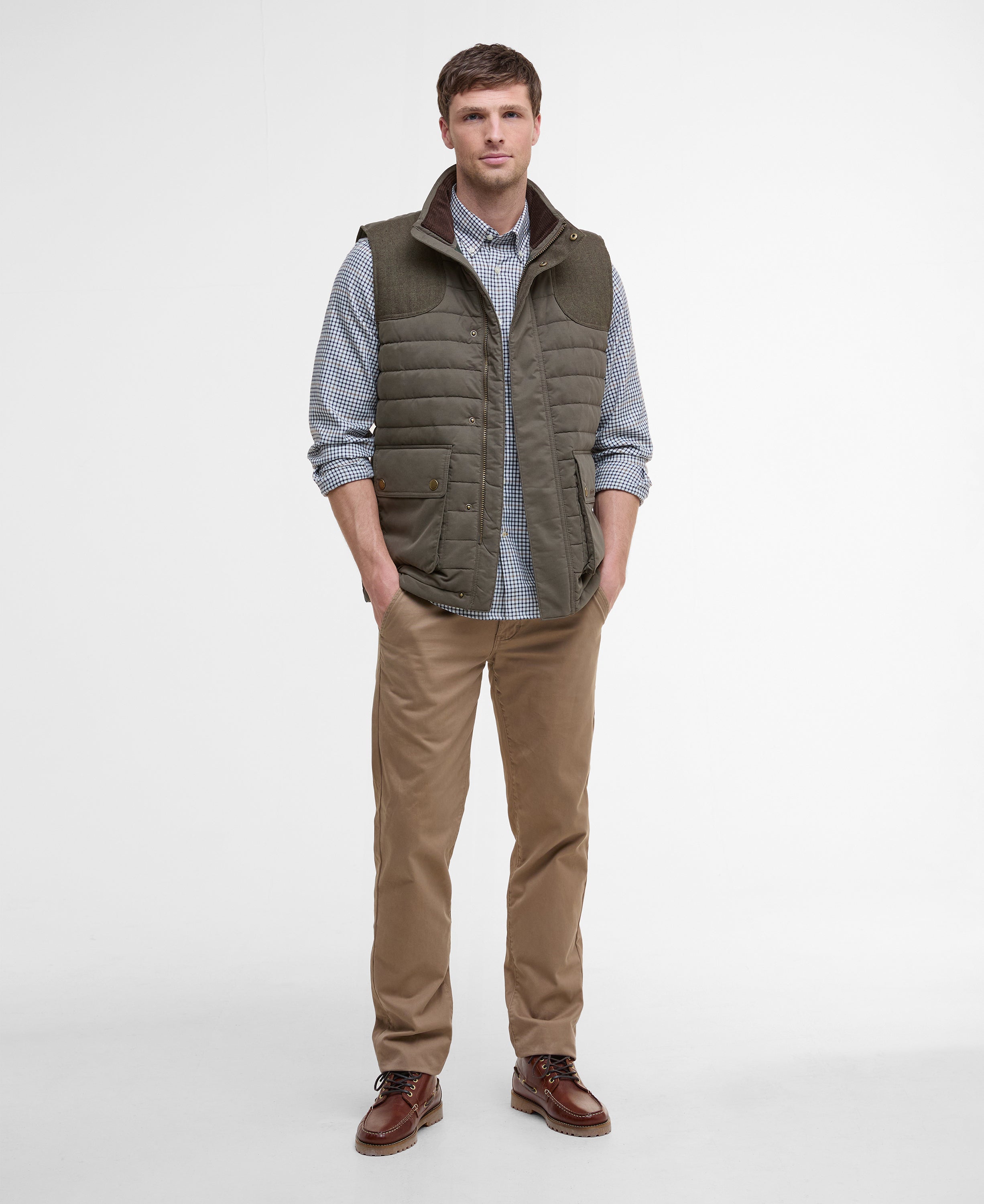 Man wearing a green puffer vest over a checkered shirt with brown pants on a white background