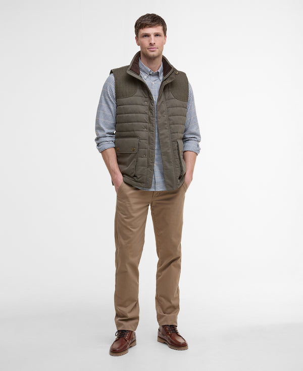 Man wearing a green puffer vest over a checkered shirt with brown pants on a white background