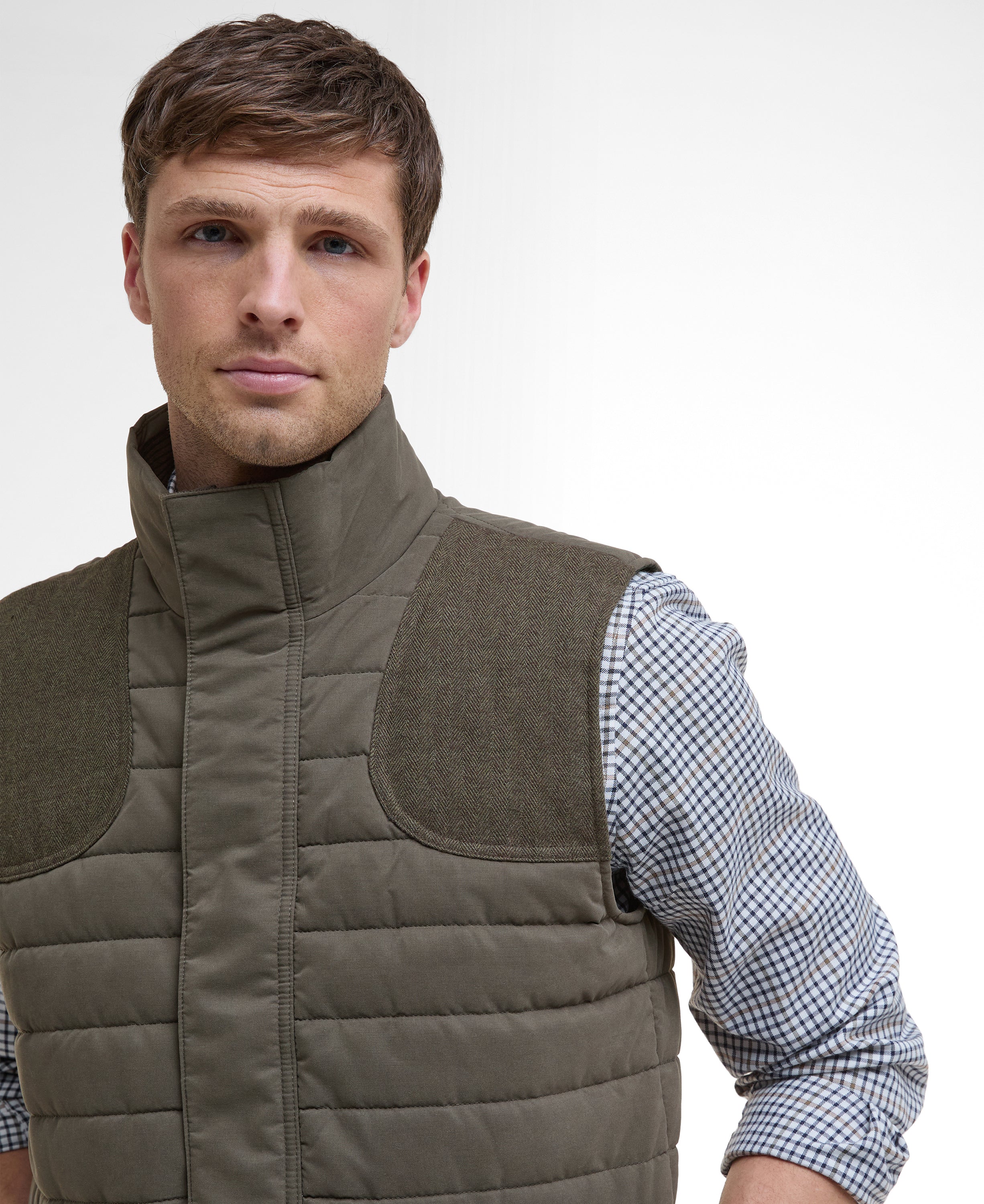 Man wearing a brown quilted vest over a checkered shirt on a white background