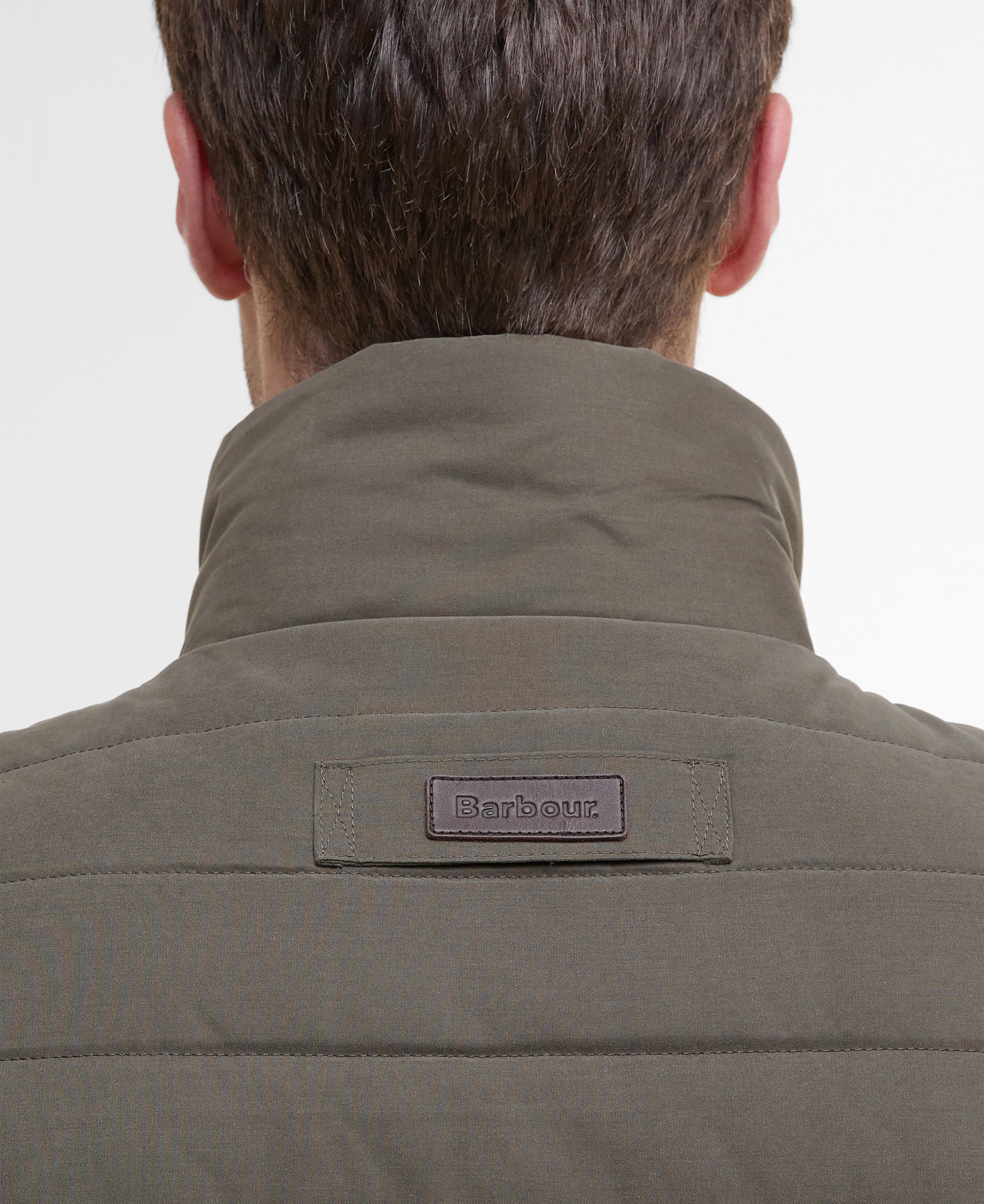 Close-up back view of a person wearing a quilted Barbour jacket in olive green, showing the high collar and the Barbour brand patch on the upper back.