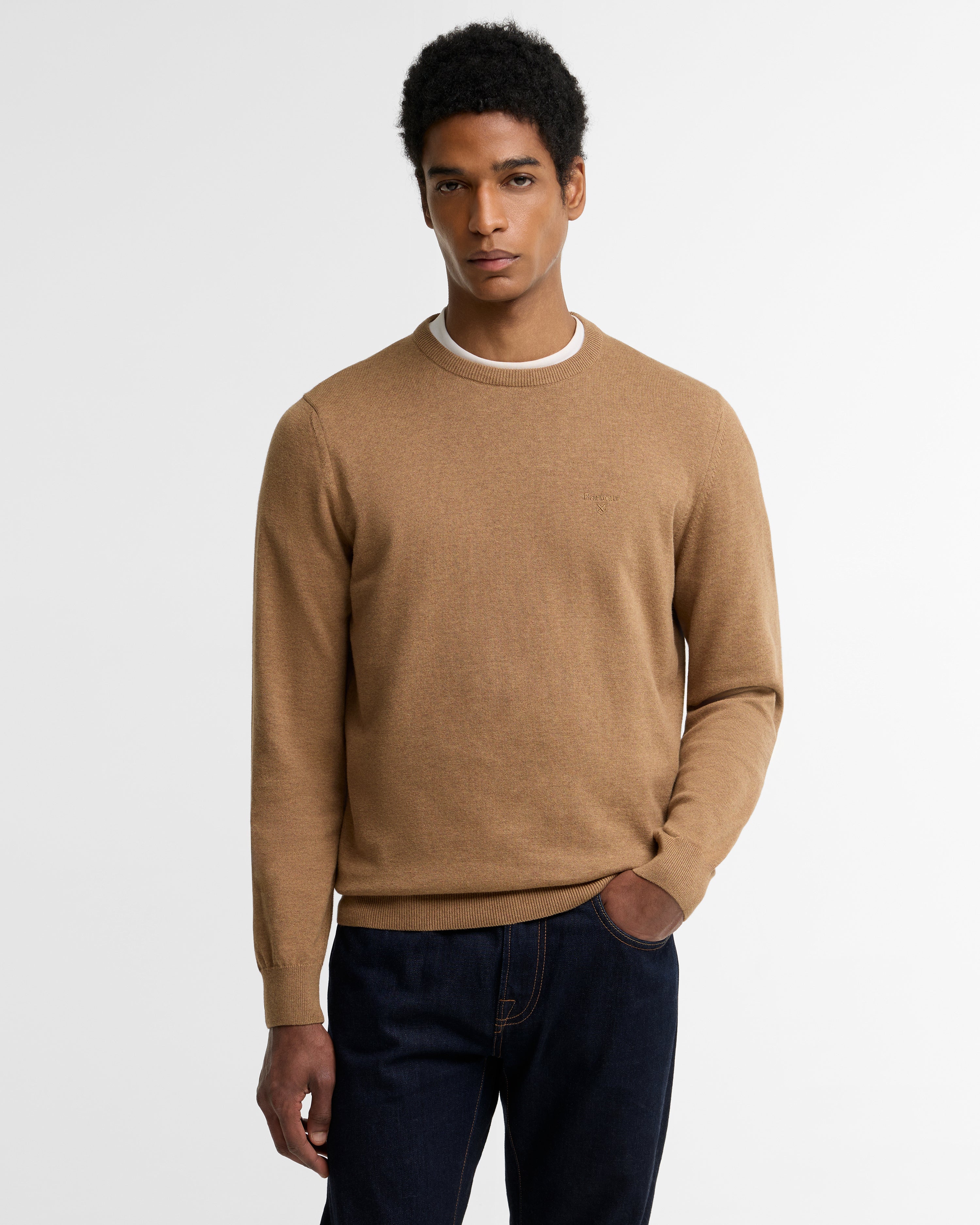 A man wearing a camel brown crewneck sweater over a white undershirt and dark blue jeans, standing against a light gray studio background.