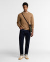 A man modeling a tan crewneck sweater, dark navy blue jeans, and beige sneakers, wearing a green crossbody bag with a black strap against a plain light gray background.