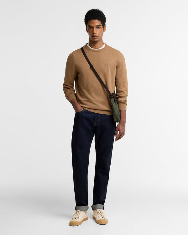 A man modeling a tan crewneck sweater, dark navy blue jeans, and beige sneakers, wearing a green crossbody bag with a black strap against a plain light gray background.