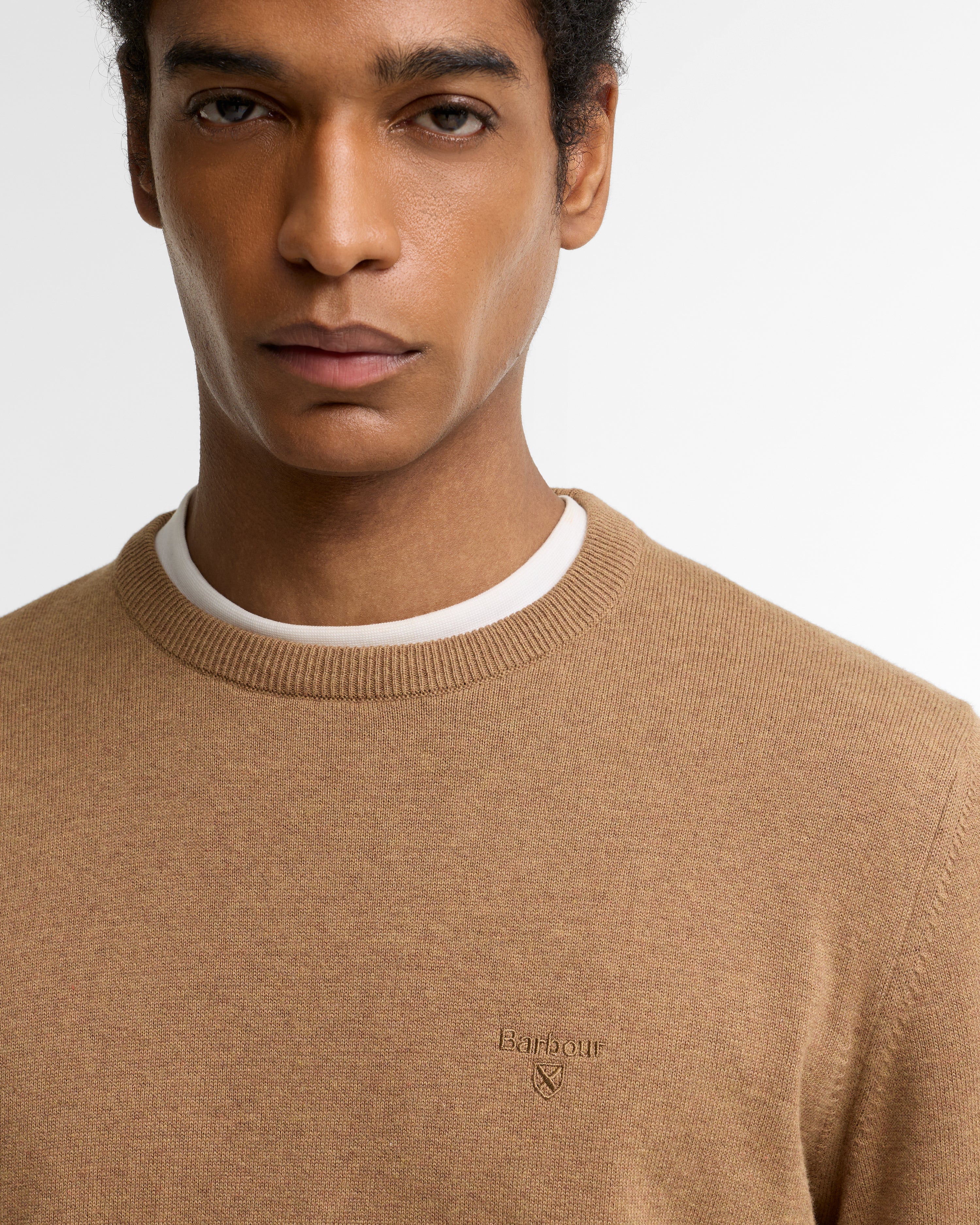 A close-up of a man wearing a light brown crewneck sweater over a white t-shirt, with a small embroidered Barbour logo on the chest.
