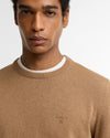 A close-up of a man wearing a light brown crewneck sweater over a white t-shirt, with a small embroidered Barbour logo on the chest.