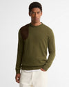 A man wearing an olive green knit sweater with dark brown suede contrast panels on the shoulders, paired with off-white trousers, against a plain white studio background.