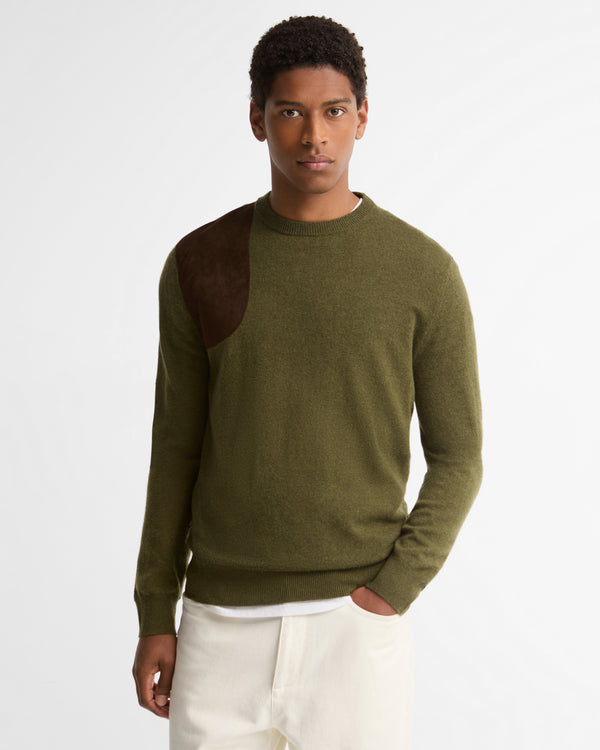 A man wearing an olive green knit sweater with dark brown suede contrast panels on the shoulders, paired with off-white trousers, against a plain white studio background.