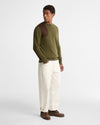 A man wearing an olive green knit sweater with brown contrast panels on the shoulders, paired with off-white trousers and brown shoes, standing against a light gray studio background.