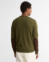 A man viewed from the back wearing an olive green crewneck sweater with dark brown elbow patches and off-white trousers, set against a plain light gray studio background.