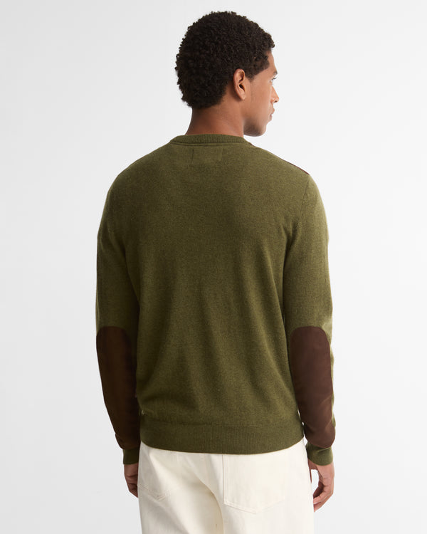 A man viewed from the back wearing an olive green crewneck sweater with dark brown elbow patches and off-white trousers, set against a plain light gray studio background.