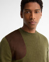 A close-up shot of a man wearing a knit crewneck sweater with an olive green body and a contrasting dark brown suede shoulder panel, set against a plain light gray background.