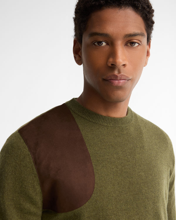 A close-up shot of a man wearing a knit crewneck sweater with an olive green body and a contrasting dark brown suede shoulder panel, set against a plain light gray background.