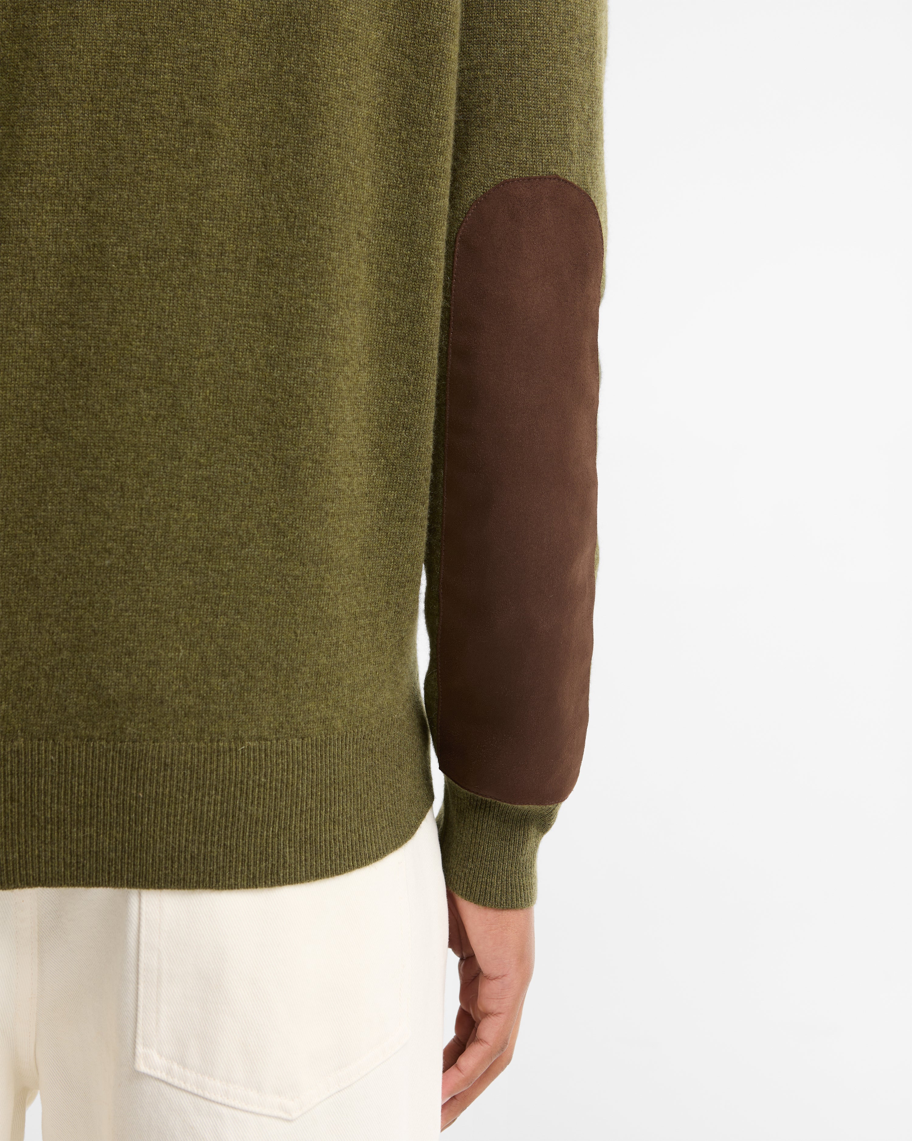 Close-up of a person wearing an olive green knit sweater with a brown suede elbow patch, paired with off-white trousers, against a plain white background.