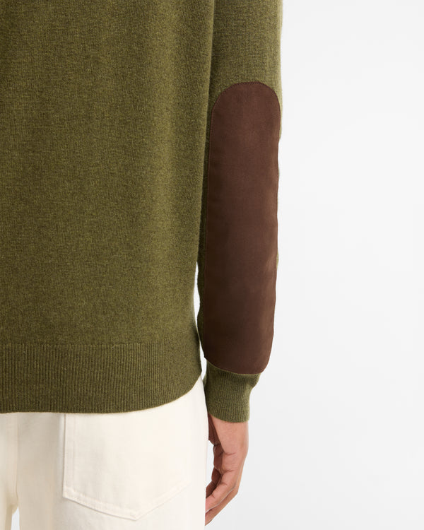 Close-up of a person wearing an olive green knit sweater with a brown suede elbow patch, paired with off-white trousers, against a plain white background.