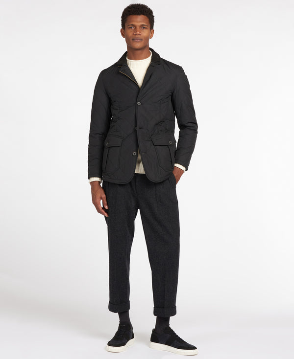 A man modeling a black utility jacket, dark charcoal trousers, a cream t-shirt, and black sneakers against a plain white studio background.
