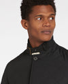 A close-up shot of a man wearing a black quilted jacket with a high stand-up collar and a button, set against a plain light gray background.