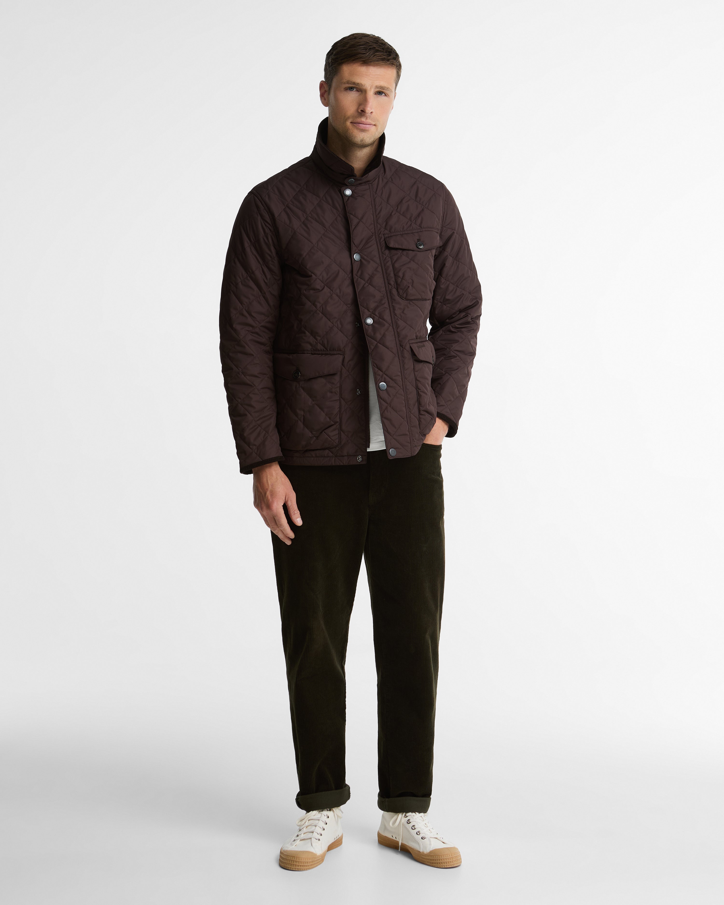 A man modeling a dark brown quilted jacket, dark brown corduroy trousers, and white sneakers against a plain light gray studio background.