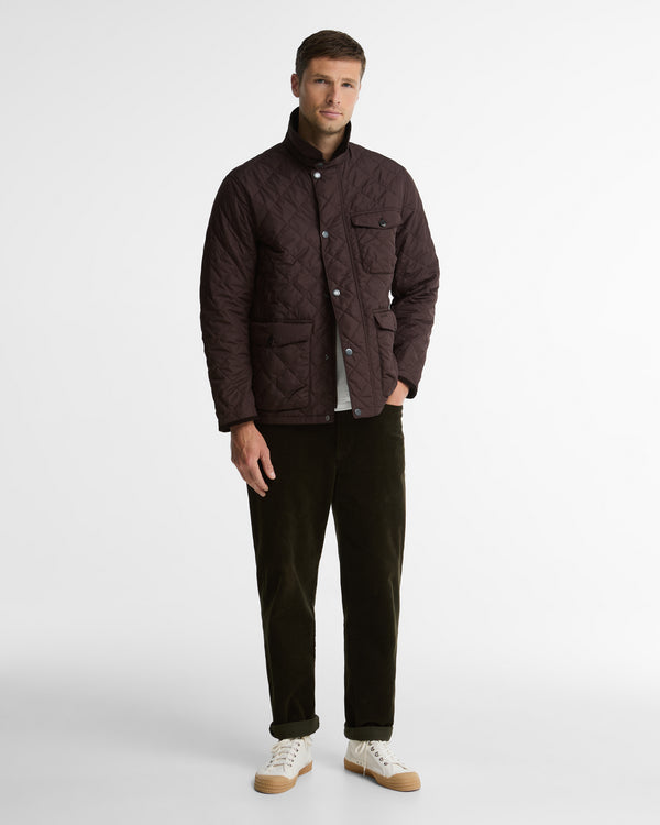 A man modeling a dark brown quilted jacket, dark brown corduroy trousers, and white sneakers against a plain light gray studio background.