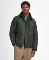 A man wearing a dark green quilted jacket with a high collar, styled over a dark shirt and beige trousers, standing against a light gray studio background.