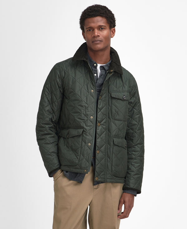 A man wearing a dark green quilted jacket with a high collar, styled over a dark shirt and beige trousers, standing against a light gray studio background.