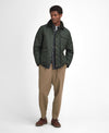 A man modeling a dark green quilted jacket over a navy shirt and tie, paired with beige trousers and brown shoes, against a plain white studio background.