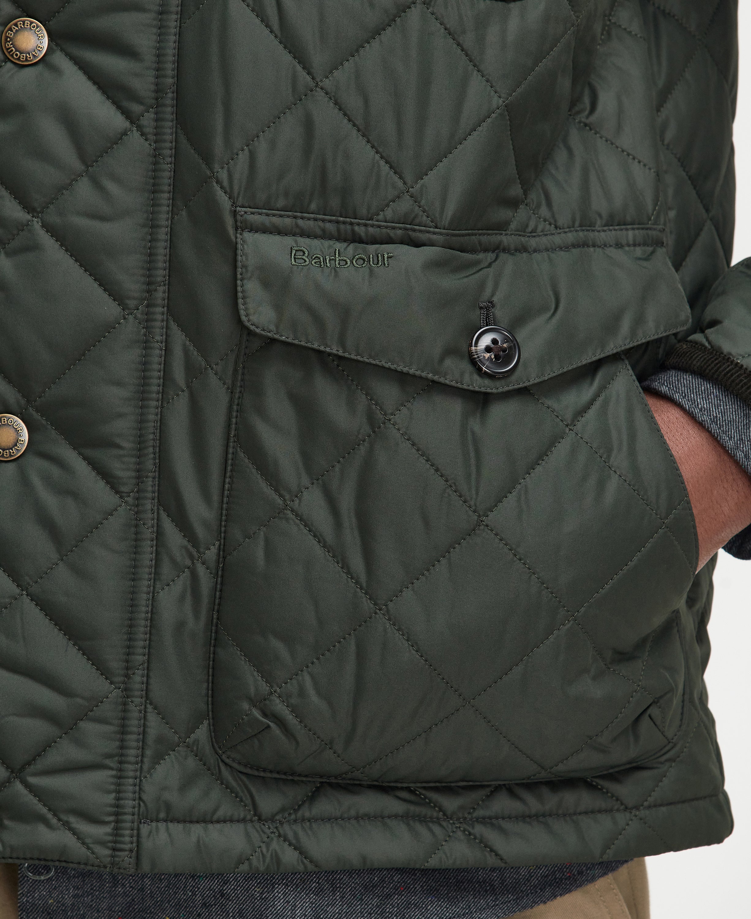 Close-up of a quilted Barbour jacket in dark olive green, showing a diamond-quilted pattern, a front flap pocket with a button, and part of a person's hand in the pocket.