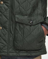 Close-up of a quilted Barbour jacket in dark olive green, showing a diamond-quilted pattern, a front flap pocket with a button, and part of a person's hand in the pocket.