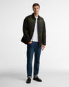 A man modeling a dark green and black plaid overshirt over a white t-shirt, paired with blue jeans and black leather shoes, against a plain light gray studio background.