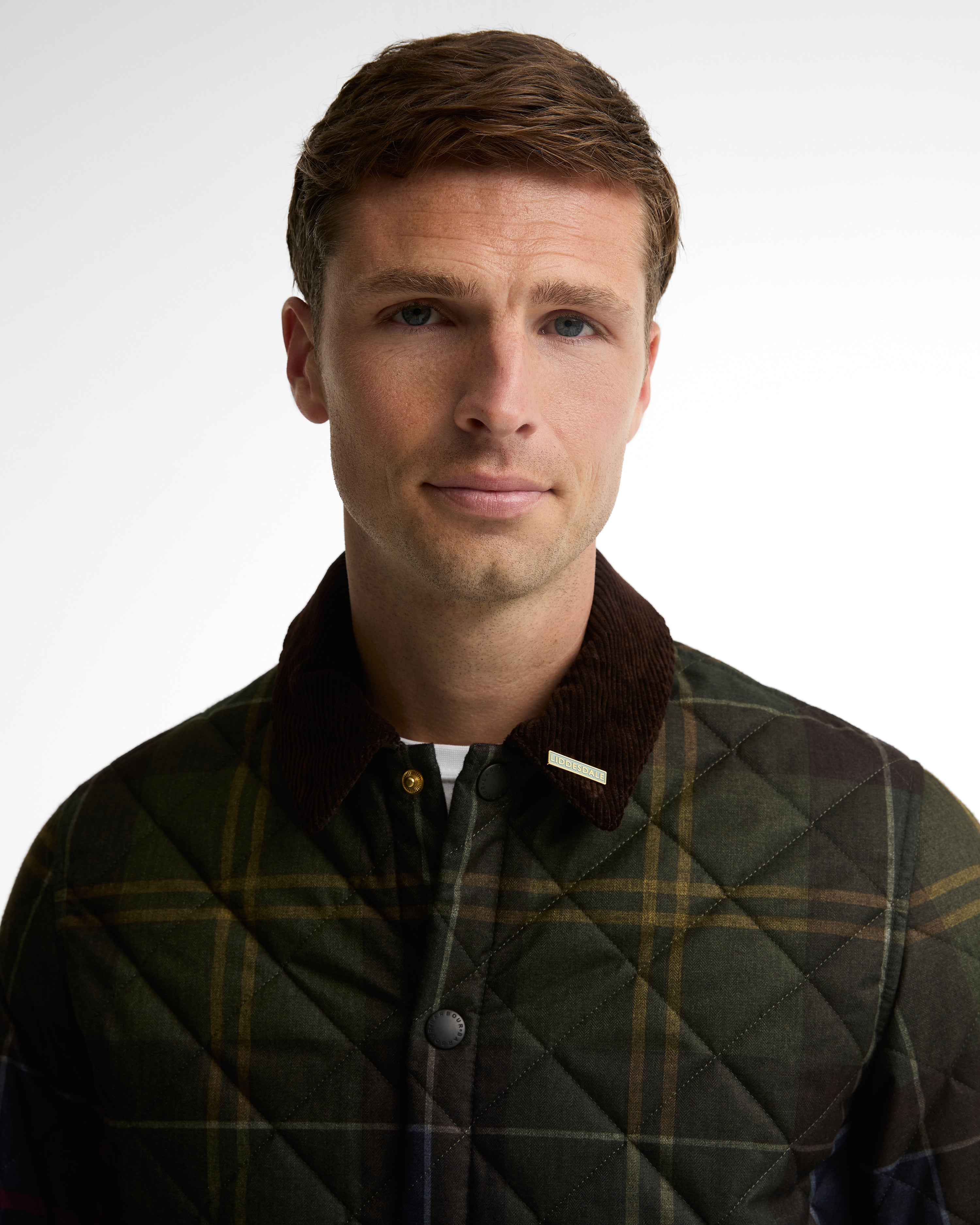 A man wearing a quilted plaid jacket with a dark brown corduroy collar, photographed against a plain light gray studio background.