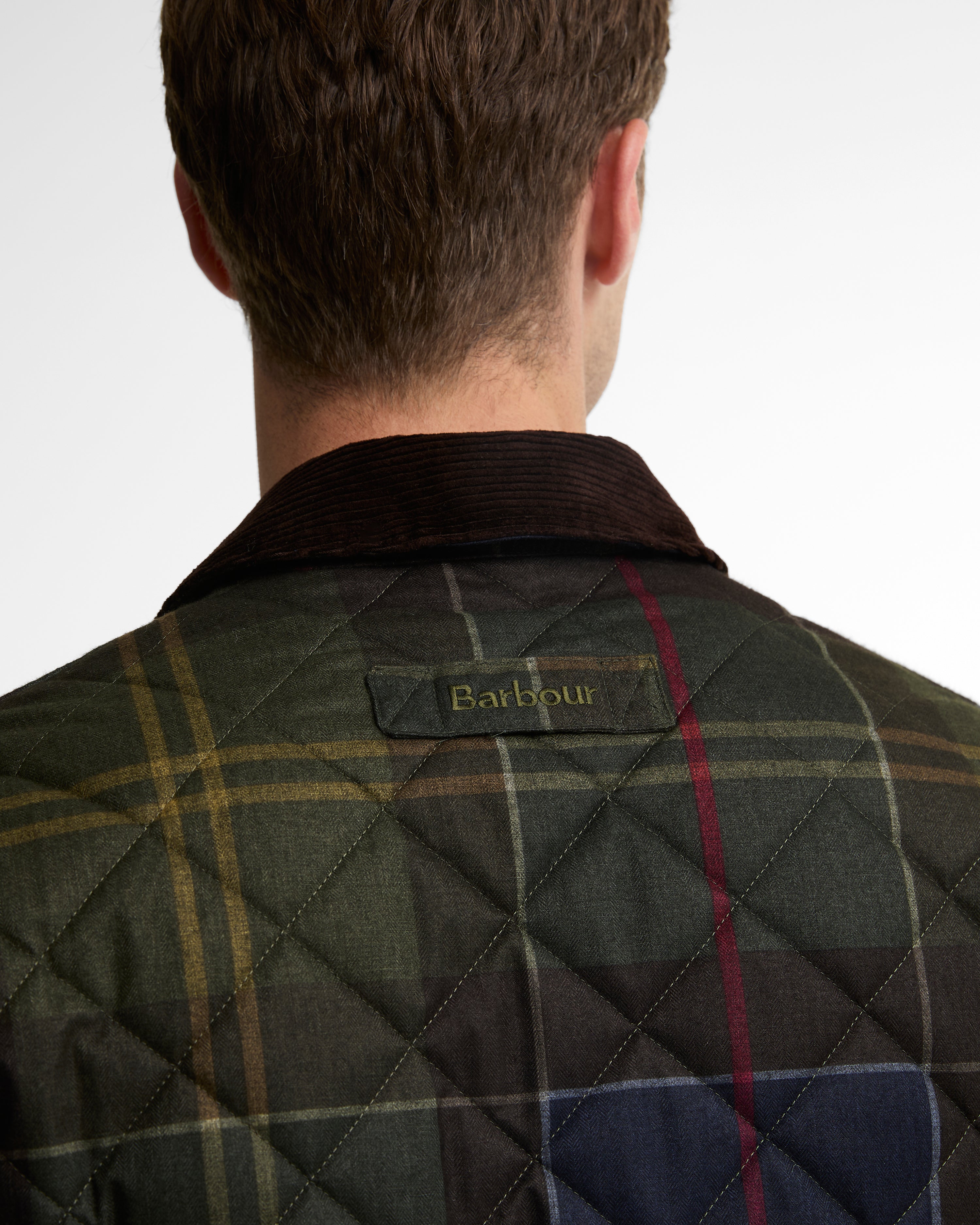 Back view of a man wearing a quilted Barbour jacket with a tartan plaid pattern in green, navy, and yellow, featuring a dark corduroy collar.