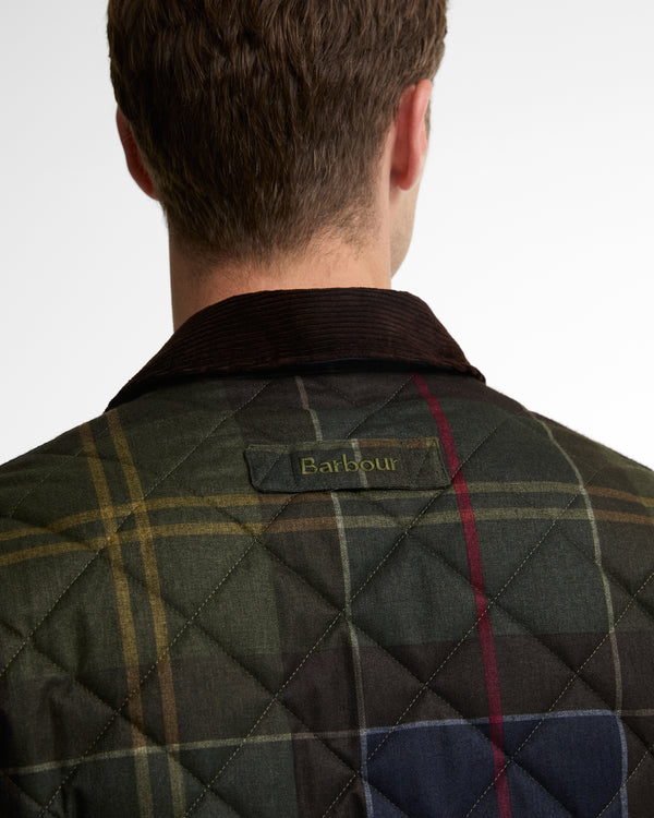 Back view of a man wearing a quilted Barbour jacket with a tartan plaid pattern in green, navy, and yellow, featuring a dark corduroy collar.