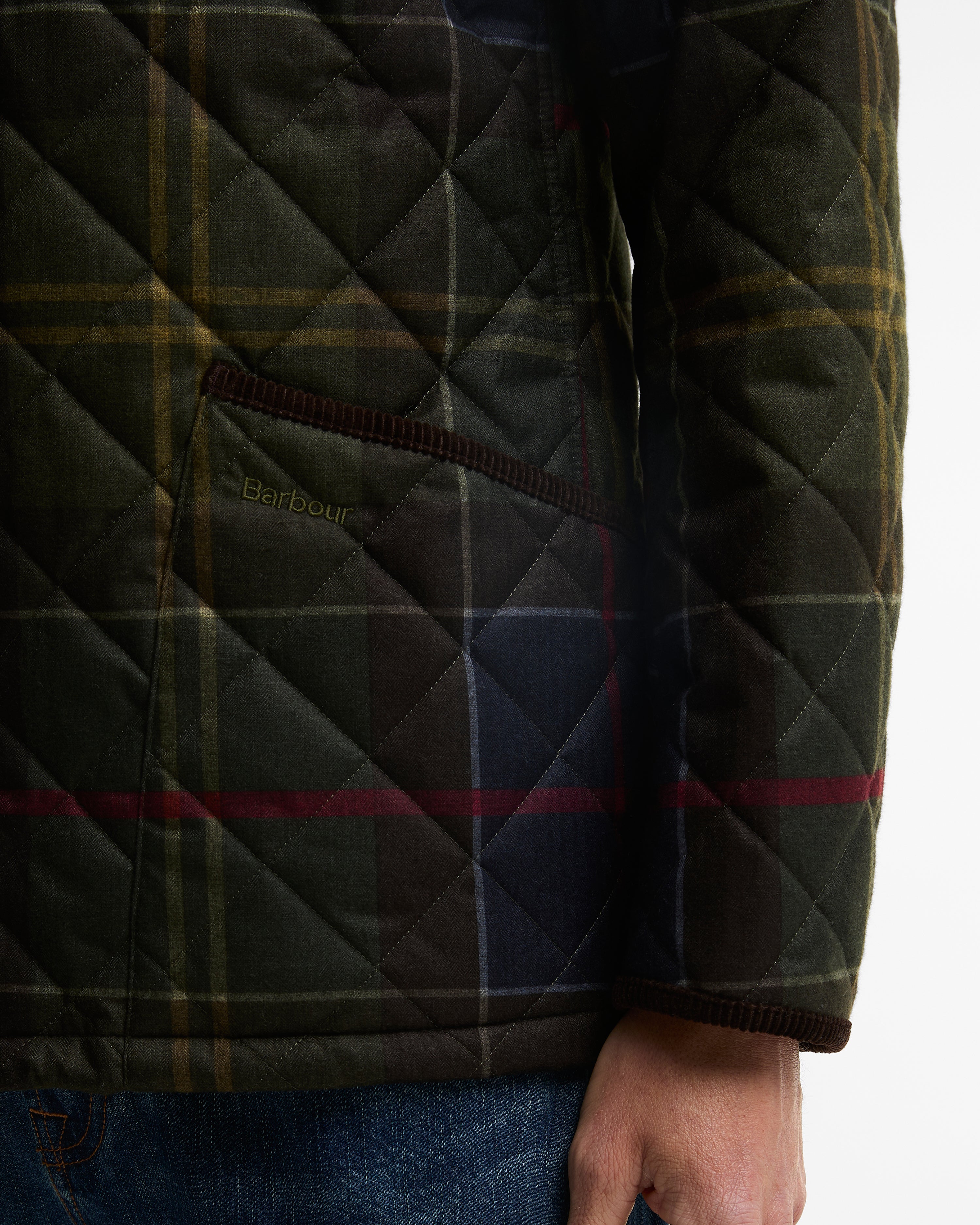 Close-up of a quilted Barbour jacket with a dark green and navy tartan pattern, featuring a diamond-quilted texture and a side pocket with the Barbour logo.
