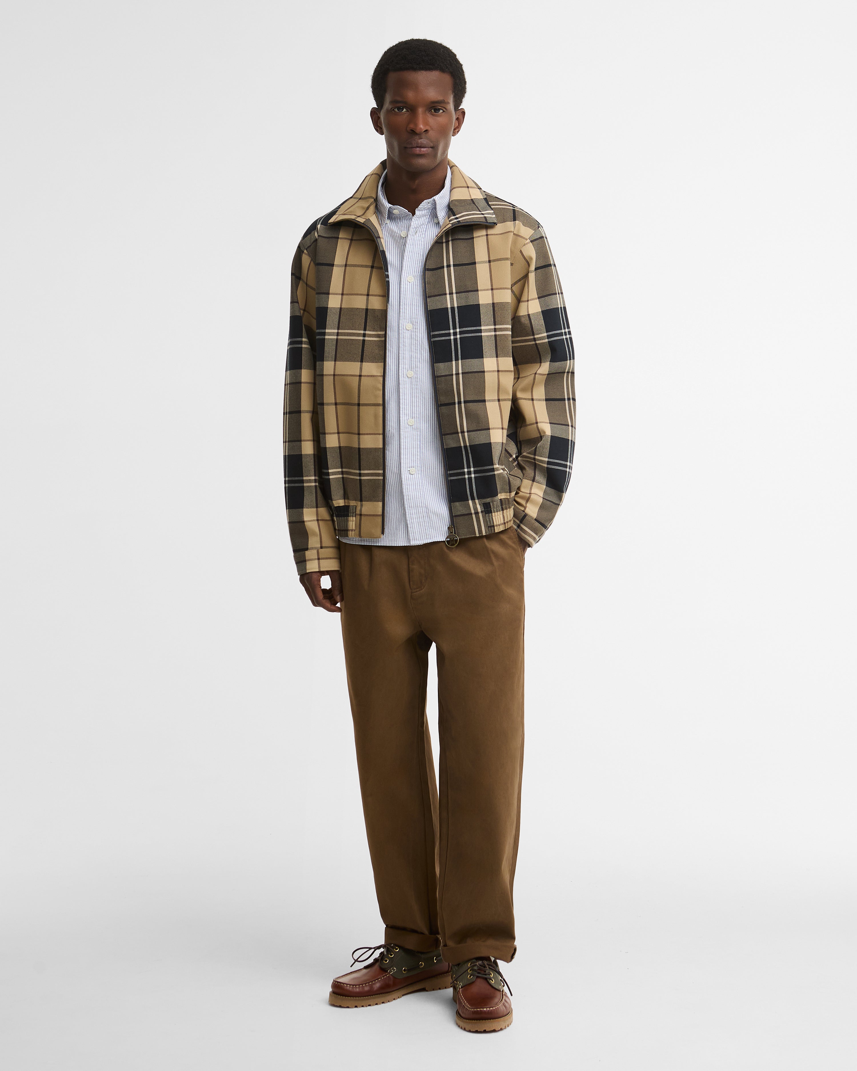 A man modeling a Barbour Tartan Showerproof Blouson Jacket over a white button-down shirt, paired with brown trousers and brown shoes.