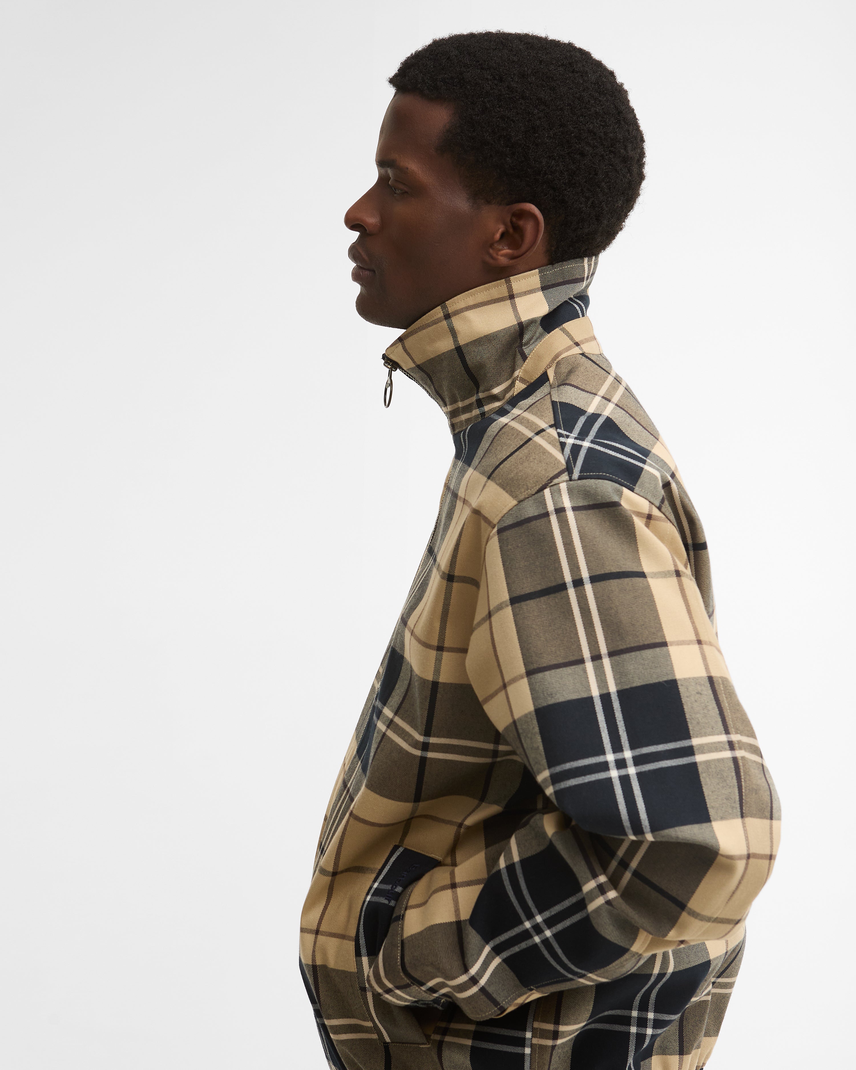 A man in profile wears a Barbour Tartan Showerproof Blouson Jacket in beige tartan, showing the stand-up collar and zip front.
