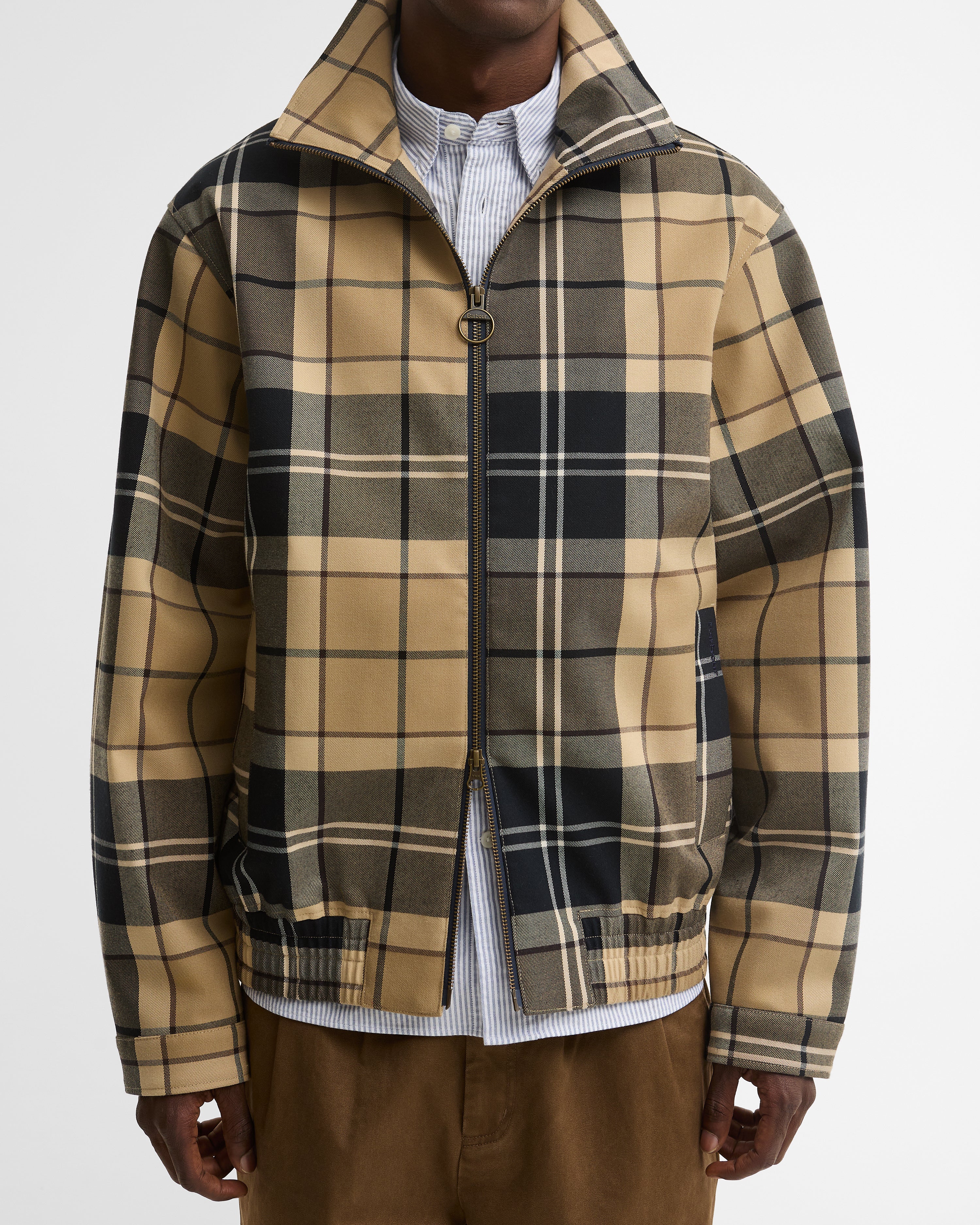 A man wearing a tan and black plaid zip-up jacket - a Barbour Tartan Showerproof Blouson Jacket - over a white striped shirt and brown pants.