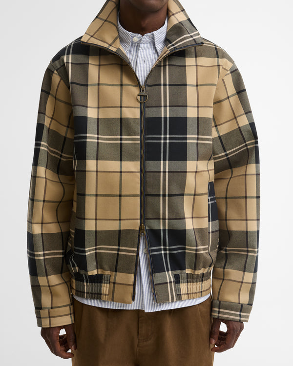 A man wearing a tan and black plaid zip-up jacket - a Barbour Tartan Showerproof Blouson Jacket - over a white striped shirt and brown pants.