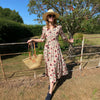Woman in a floral dress and sun hat standing by a fence in a garden.
