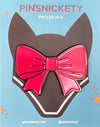 A glossy enamel bow shaped pin with a large hot pink bow, displayed on its dark brown backing card against a light blue background with decorative text and sparkles.