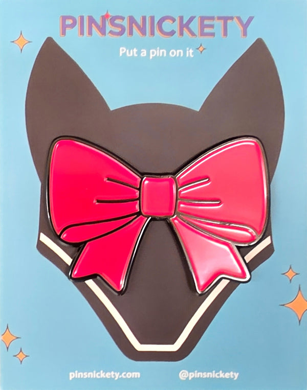 A glossy enamel bow shaped pin with a large hot pink bow, displayed on its dark brown backing card against a light blue background with decorative text and sparkles.